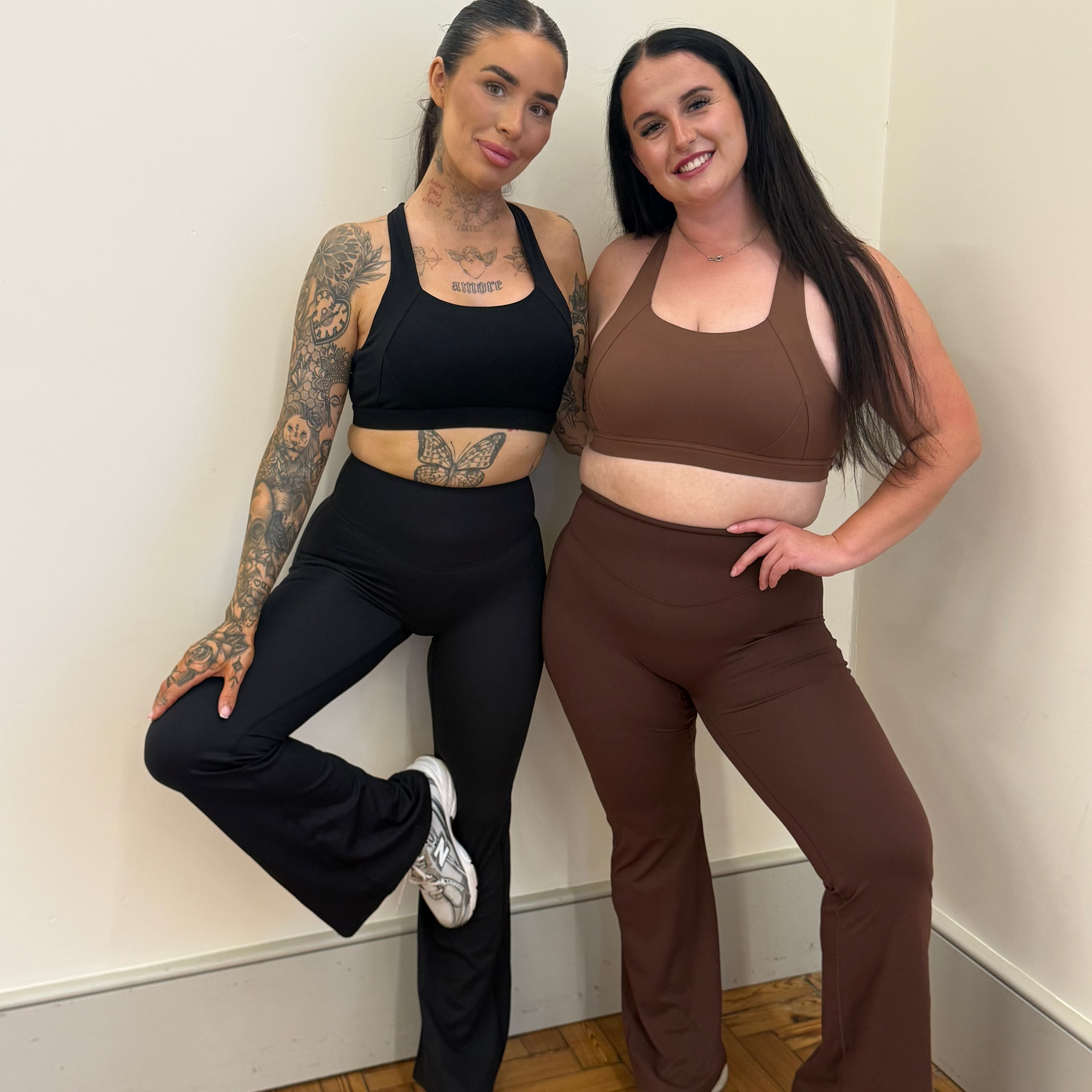 Two women posing together in matching athletic sets, one in a black set and the other in a brown set, standing in a corner of a room with a wooden floor.